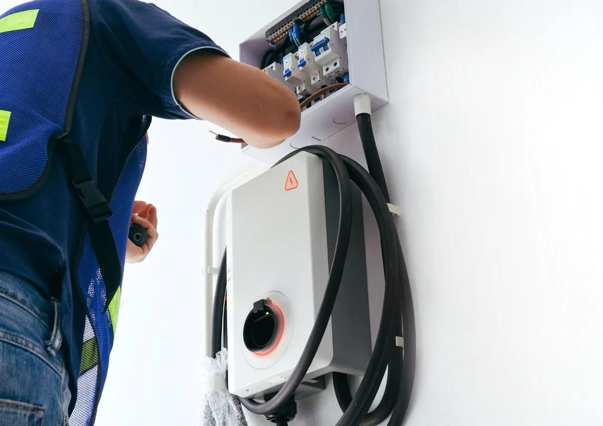 Electrician in a safety vest wiring a small breaker box above a wall-mounted EV charger with its cable coiled
